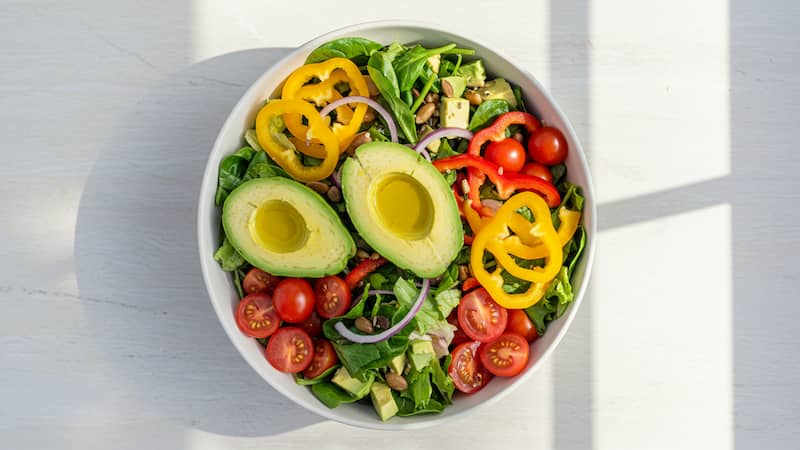 A large, vibrant salad in a white bowl, filled with mixed greens, avocado, and bell peppers.